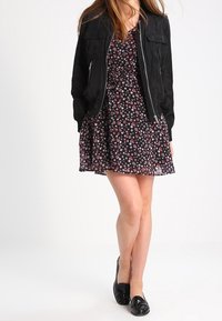 Mujer vestida con un vestido floral negro acompañado de una chaqueta negra con cremallera y mocasines negros, de pie con las piernas cruzadas frente a un fondo blanco.