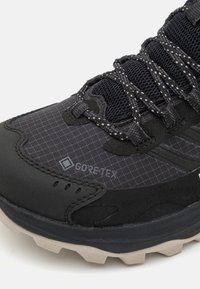 Czarne buty trekkingowe z logo Gore-Tex, z fakturowaną powierzchnią i siateczkowym materiałem, wzmocnionym noskiem oraz jasnoszarym, gumowym podbiciem z wypustkami zapewniającymi dodatkową przyczepność.
