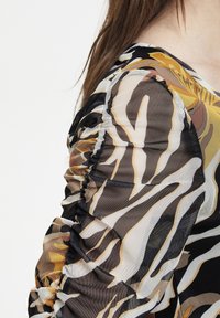 Top trasparente con dettagli arricciati, presenta un motivo astratto nero, marrone e oro. Tessuto morbido con una texture leggera.