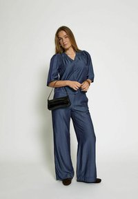 Mornarsko modra svila jumpsuit z rokavi do tri četrtine, v-izrezom in gumbom spredaj; v kombinaciji s slim črno torbico. Hlače so širokega kroja.
