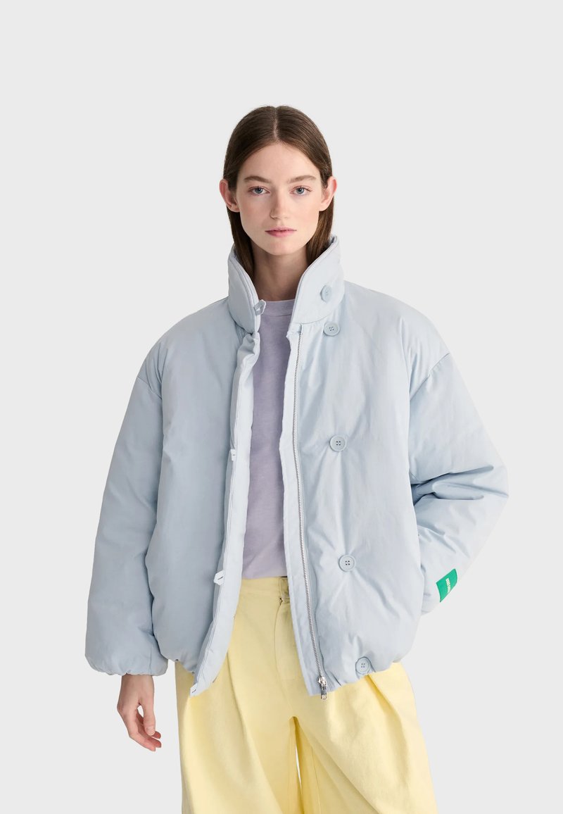 Jeune femme portant une doudoune bleu clair par-dessus un haut lavande et un pantalon jaune, debout devant un fond uni clair.
