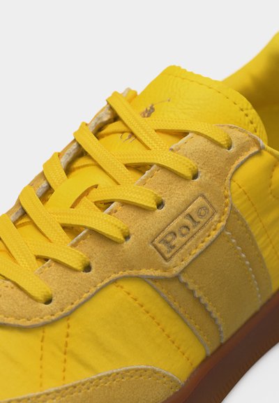 Κίτρινο αθλητικό παπούτσι με πάνελ από γούνα και ύφασμα, κίτρινα κορδόνια, ανάγλυφο λογότυπο Polo στο πλάι και καφέ λαστιχένια σόλα.