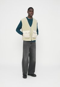 Gilet in pile giallo pallido con zip frontale e due grandi tasche crema. Abbinato a una felpa color teal e jeans grigi a gamba larga. Scarpe nere.