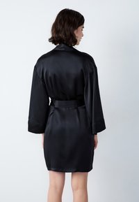 Robe di raso nero con vita a cintura, dotata di maniche lunghe e una texture liscia. Mostrata da dietro, evidenziando un design minimale.
