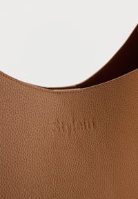 Κοντινό πλάνο καφέ δερμάτινης τσάντας με υφή, με κυρτή πάνω άκρη και διακριτικό ανάγλυφο όνομα εμπορικού σήματος "Stylein".