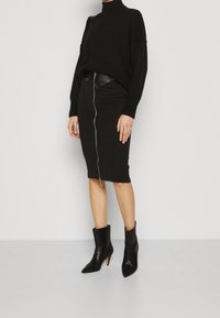 Suéter negro de punto con un cuello alto, combinado con una falda lápiz negra con una cremallera frontal. Botines de cuero negro con puntera afilada.