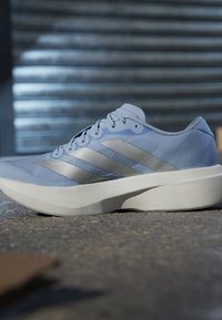 Jasnoniebieski but sportowy z białą podeszwą i metalicznie srebrnymi trzema paskami po bokach, spoczywający na nierównej powierzchni ziemi.