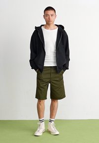 Fato com fecho éclair preto, camiseta branca canelada, calções verde-oliva e ténis de cano alto bege com meias listradas em preto e branco. Fundo liso.
