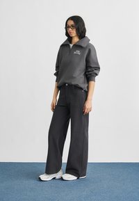 Γυναίκα με μαύρα γυαλιά, σκούρο γκρι φούτερ New Balance με φερμουάρ μέχρι τη μέση, φαρδιά μαύρη παντελόνα και λευκά αθλητικά παπούτσια σε μπλε χαλί.