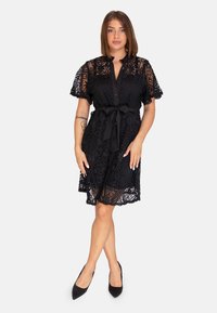 Robe en dentelle noire avec des manches courtes, fermeture boutonnée à l'avant et taille cintrée. Présente des motifs élaborés et une jupe évasée. Portée avec des talons noirs.