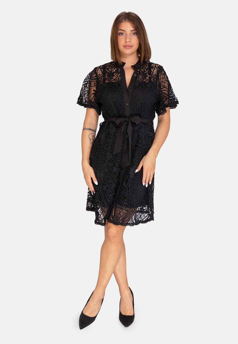 Robe en dentelle noire avec des manches courtes, fermeture boutonnée à l'avant et taille cintrée. Présente des motifs élaborés et une jupe évasée. Portée avec des talons noirs.