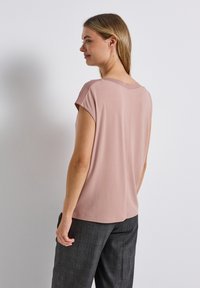 Top rosa a maniche corte realizzato in tessuto morbido. Collo rotondo, vestibilità comoda e dettagli di cucitura sottili sulle spalle. Indossato con pantaloni scuri a fantasia.