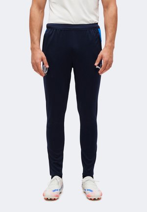 Homme portant un pantalon de sport slim bleu marine avec logo Puma blanc et des crampons blancs avec des touches multicolores, se tenant devant un fond uni.