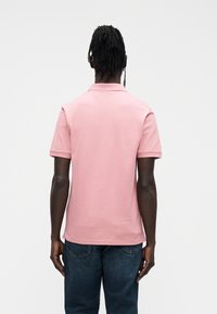 Vyriški trumpomis rankovėmis rožinės spalvos polo marškinėliai iš švelnaus medvilnės audinio, su klasikiniu apykakle ir lygiu paviršiumi, derinami su tamsiai mėlynais džinsiniais.