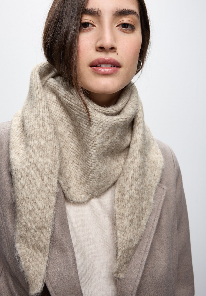 Écharpe en tricot beige avec une texture douce, présentant un design drapé. Portée sur un haut clair, partiellement visible sous un manteau pâle.