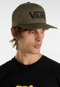 Gorra verde de tela con una forma estructurada, que presenta un destacado logo negro de "VANS" en la parte frontal y un ala plana.