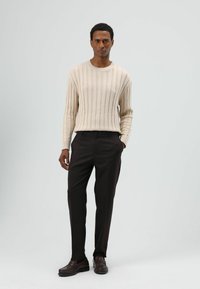 Beige geribbelde gebreide trui met lange mouwen, gecombineerd met donkerbruine rechte broek en bijpassende bruine loafers, tegen een lichte achtergrond.