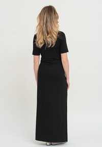 Schwarzes Maxikleid aus weichem Stoff. Kurze Ärmel, runder Ausschnitt und seitlicher Schlitz. Das Model steht mit dem Rücken zur Kamera, die langen, welligen Haare sind sichtbar.