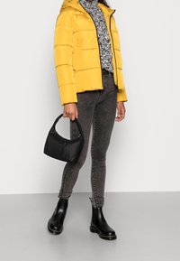 Hřejivá žlutá bunda s kapucí, šedý vzorovaný svetr, tmavé džíny, černé kotníkové boty a malá černá taška s kulatou rukojetí.