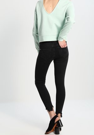 Person trägt schwarze enge Jeans mit ausgefransten Säumen, hellgrünes langärmeliges Oberteil mit tiefem Rückenausschnitt und schwarze Stiefeletten mit Absatz.