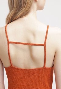 Žena s bledou pletí a světlými vlasy, která má na sobě bezramínkové ribbené oranžové tričko s tenkými ramínky a horizontálním popruhem na zádech.