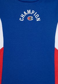 T-shirt en coton bleu avec des panneaux latéraux rouges et blancs, ornée du logo "CHAMPION" imprimé en blanc en arc sur la poitrine. Texture lisse.