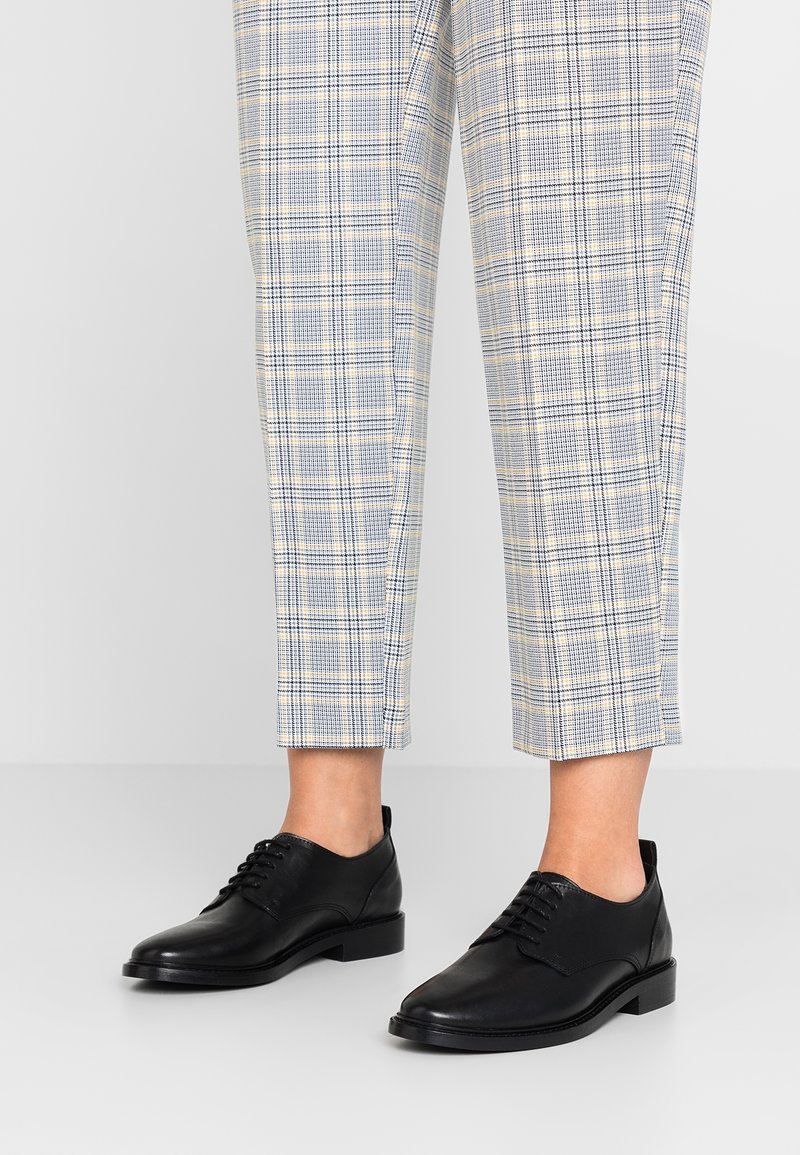 Pantalon à carreaux longueur cheville associé à des chaussures habillées noires en cuir à lacets sur un fond neutre.