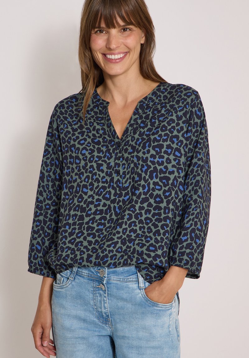 Femme souriante portant un chemisier à manches longues bleu foncé et vert à motif léopard et un jean bleu clair, une main dans la poche.
