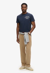 Marineblaue Baumwoll-T-Shirt mit weißem Drucklogo, beigefarbene Baumwollhose, grauer Pullover um die Taille gebunden und weiße Turnschuhe.