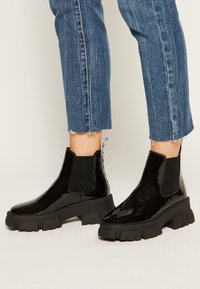 Bottines Chelsea en cuir verni noir avec panneaux latéraux en élastique côtelé et semelle en caoutchouc épaisse. Équipées d'une languette au talon pour un enfilage facile.