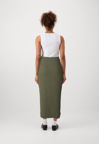 Fustă maxi tricotată de culoare verde măslin, combinată cu un top alb fără mâneci; prezintă un talie ajustată și o textură netedă, purtată cu balerini negri și șosete albe.