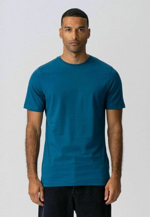 Mees lühikeste juustega, kandes lihtsat türkiissinist ümmarguse kaelusega lühikese varrukaga T-särki, seisab neutraalse halli tausta ees.