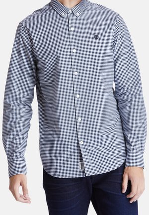 Mann trägt langärmeliges, marineblau-weiß kariertes Hemd mit Knöpfen und dunkelblaue Jeans, steht vor neutralem Hintergrund.