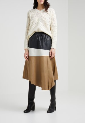 Un pull en tricot crème avec un col en V, associé à une jupe en cuir bicolore noire, blanche et beige. De grandes bottes à talons noires.