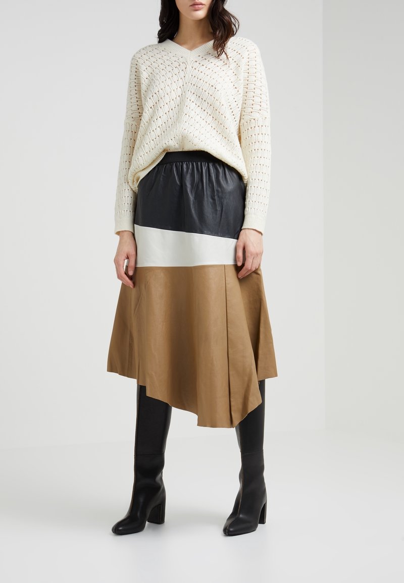 Maglione lavorato a maglia color crema con scollatura a V, abbinato a una gonna in pelle a blocchi di colore nei toni nero, bianco e beige. Stivali alti neri con tacco.