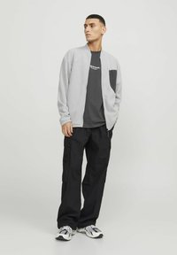 Giacca zip grigia chiara con tasca sul petto nera, t-shirt nera sotto e pantaloni cargo neri, indossati con scarpe da ginnastica.