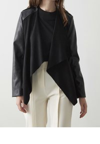 Veste en cuir noir au design drapé, dotée d'un col et de manches longues, associée à un pantalon crème taille haute. Texture lisse.