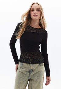 Schwarzes Langarmoberteil mit einer Spitzen-Oberpartie und unterem Saum, das ein körperbetontes Design und eine weiche Textur aufweist, kombiniert mit hellen Denim-Hosen.