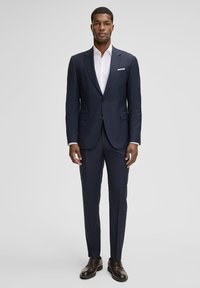 Marineblauer, maßgeschneiderter Anzug aus glattem Stoff, bestehend aus einer Ein-Knopf-Jacke, schlanken Hosen und einem kontrastierenden weißen Hemd. Dunkle Herrenschuhe vervollständigen den Look.