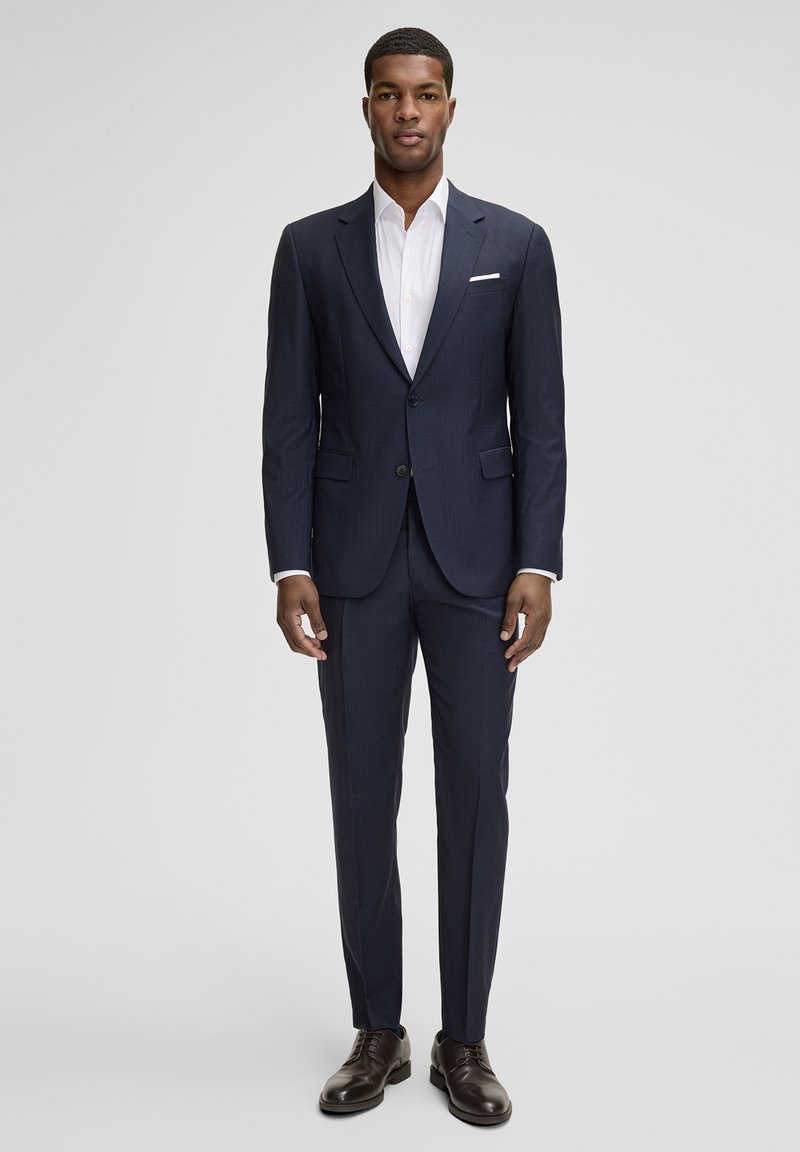 Marineblauer, maßgeschneiderter Anzug aus glattem Stoff, bestehend aus einer Ein-Knopf-Jacke, schlanken Hosen und einem kontrastierenden weißen Hemd. Dunkle Herrenschuhe vervollständigen den Look.