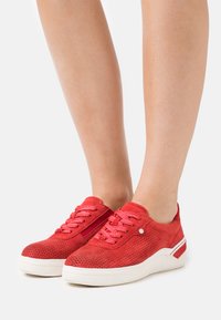 Scarpe da ginnastica rosse con un design perforato, dotate di lacci rossi accesi, suola bianca e una finitura testurizzata, mostrate su gambe nude.