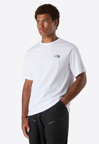 Mladý muž ve bílém tričku The North Face a černých kalhotách, stojící s jednou rukou v kapse, mírně se otáčející doleva.