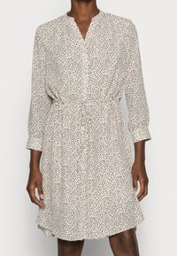 Langarm-Blusekleid aus cremefarbenem Stoff mit kleinen schwarzen, verstreuten Punkten, ausgestattet mit einem taillierten Gürtel und einem leicht ausgestellten Saum.