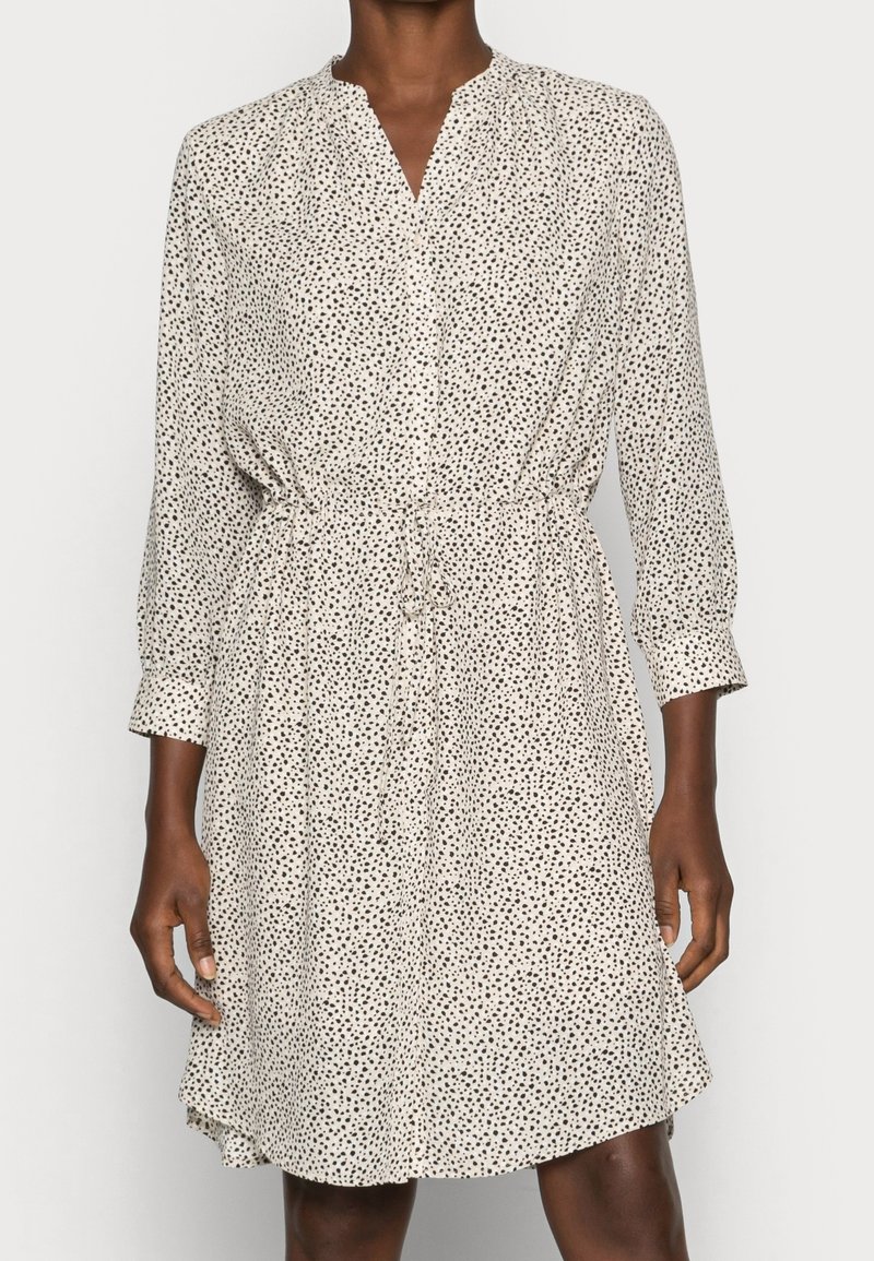 Langarm-Blusekleid aus cremefarbenem Stoff mit kleinen schwarzen, verstreuten Punkten, ausgestattet mit einem taillierten Gürtel und einem leicht ausgestellten Saum.