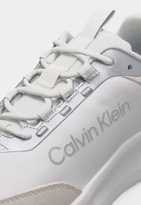 Άσπρο αθλητικό παπούτσι Calvin Klein με άσπρα κορδόνια, ασημένιες τρύπες και χοντρή σόλα, εμφανιζόμενο σε κοντινή λήψη.