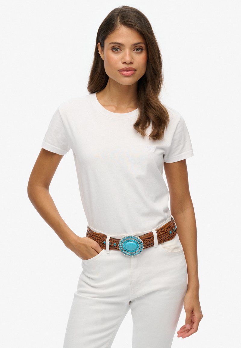 Camiseta blanca de algodón con mangas cortas, combinada con un cinturón de fibra trenzada marrón que presenta una hebilla ovalada turquesa y pequeños acentos azules.