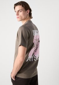 Olive green short-sleeve t-shirt with a graphic print in pink and white on the back, featuring design elements resembling abstract shapes.