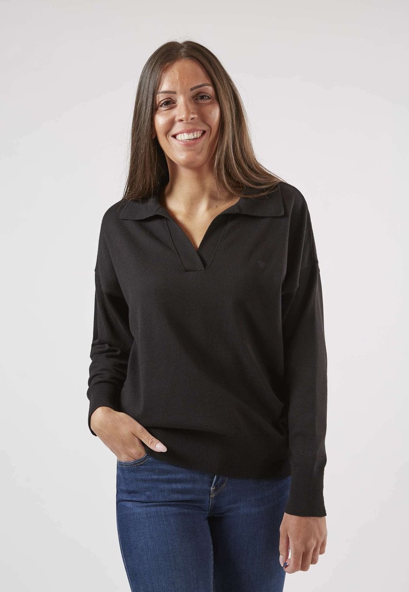 Pull noir avec col et décolleté en V. Manches longues, poignets côtelés et petit logo en forme de cœur sur le côté gauche. Jean visible.