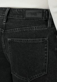 Vaqueros de mezclilla negra con un acabado texturizado, que presentan una etiqueta de la marca YAS en la cinturilla y un bolsillo trasero con detalles de costura sutiles.