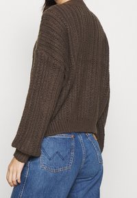 Brązowy sweter z dzianiny o fakturowanym wzorze, luźnym kroju i prążkowanym dole. W zestawie z niebieskimi dżinsami z przeszyciami na tylnych kieszeniach.